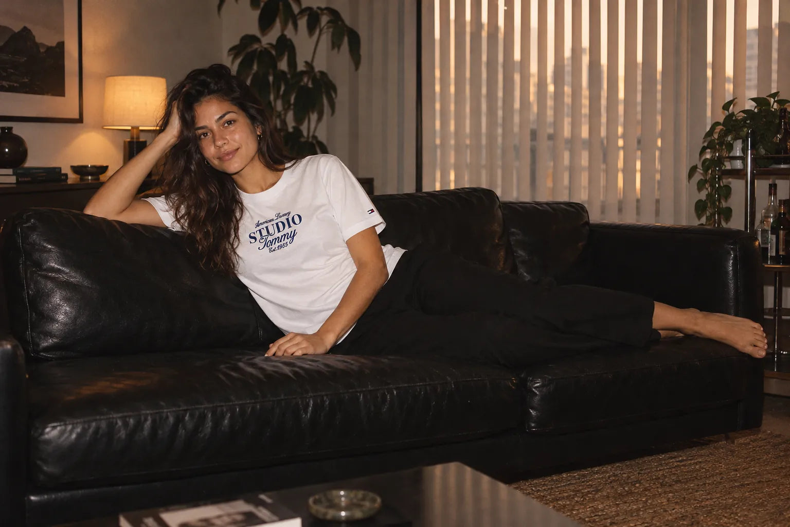 Mulher sorrindo, deitada em um sofá de couro preto, de camiseta branca Studio Tommy e calça preta