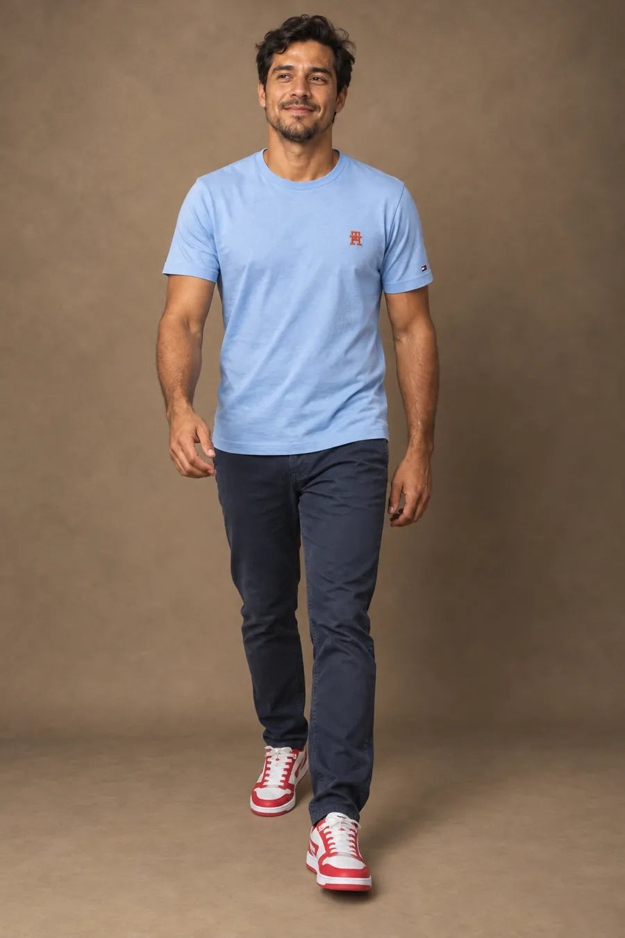 Homem caminhando de camiseta azul clara, calça azul-marinho e tênis branco e vermelho, em estúdio