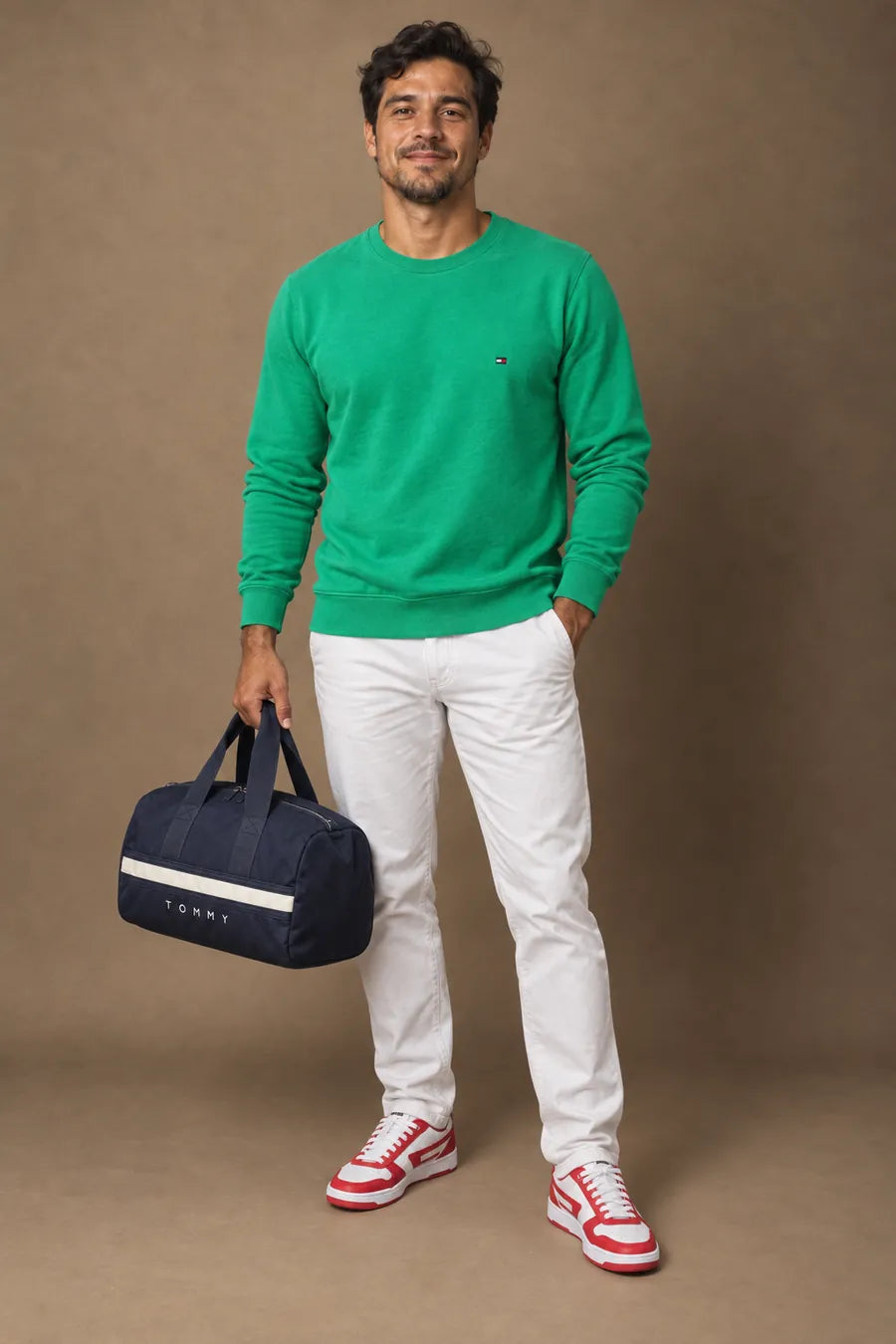 Homem de moletom verde, calça de sarja clara, tênis branco e vermelho e bolsa duffle azul-marinho, em estúdio
